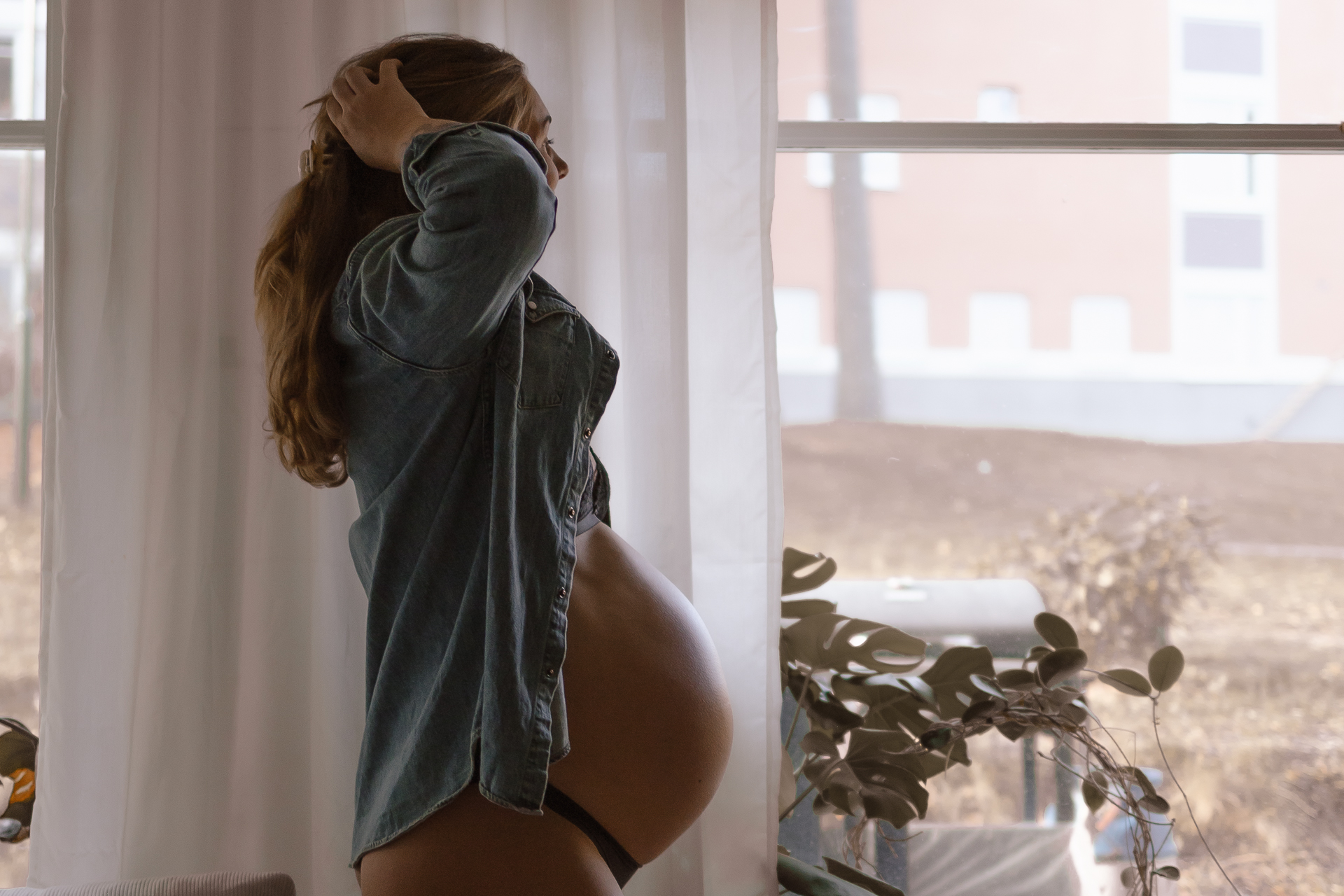 Gravid kvinna står vid fönstret med händerna i håret.